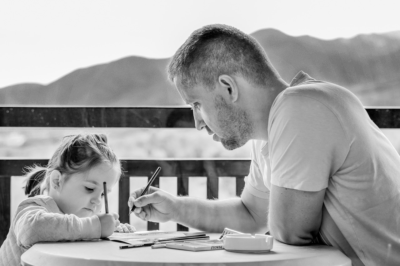 dad and 3 year old sitting at a table outside on a patio. The dad is helping his daughter with writing.